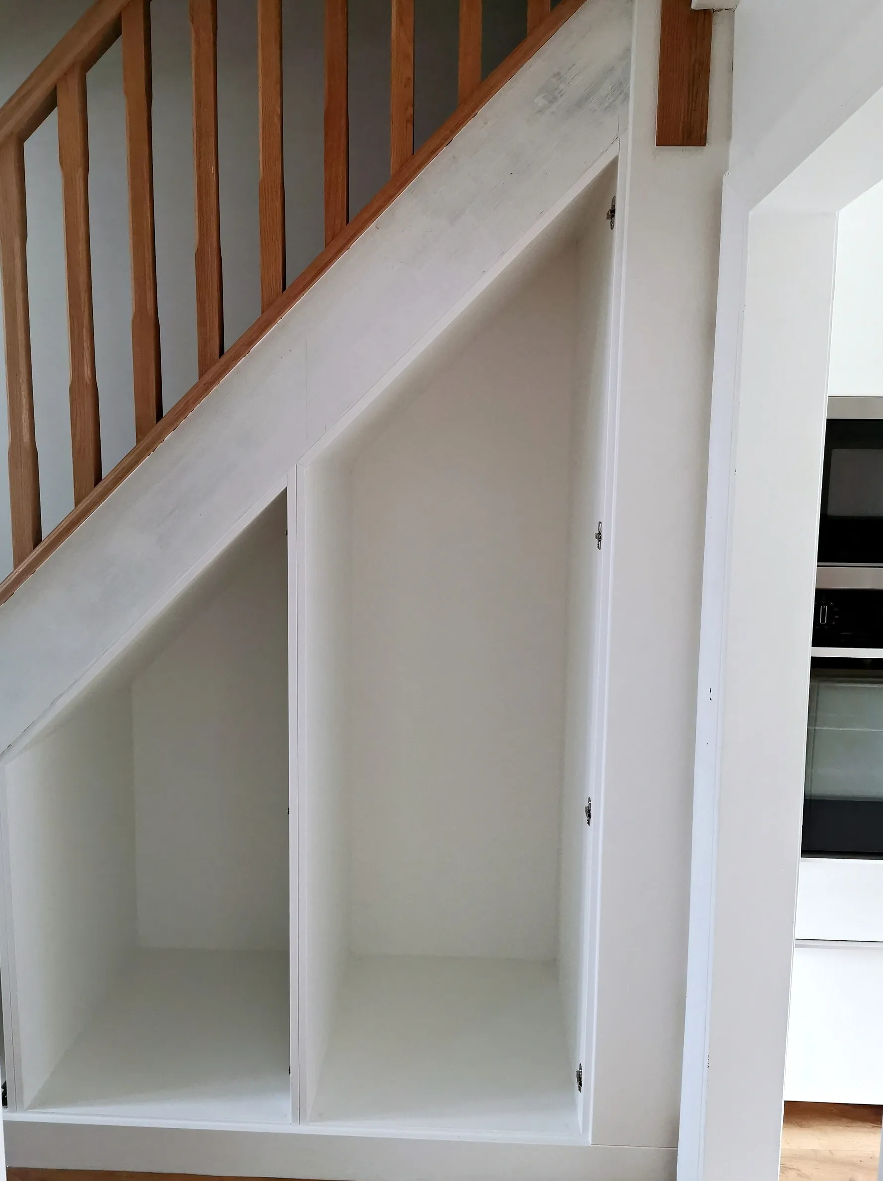 Three white under stairs storage carcasses fixed in place beneath an oak balustrade staircase, before the doors are fitted