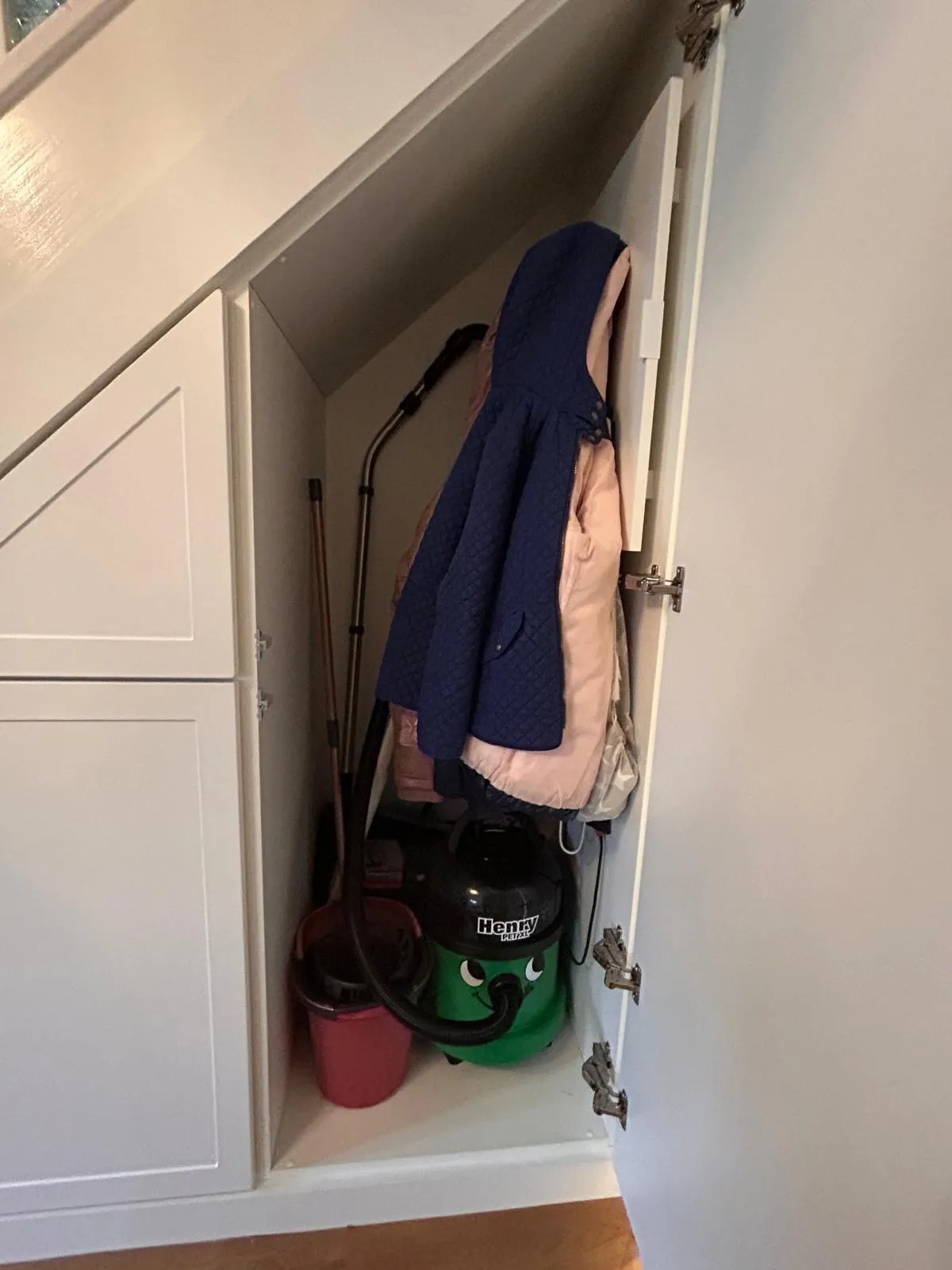 A tall under stairs cupboard open with a vacuum, mop and bucket below hanging coats