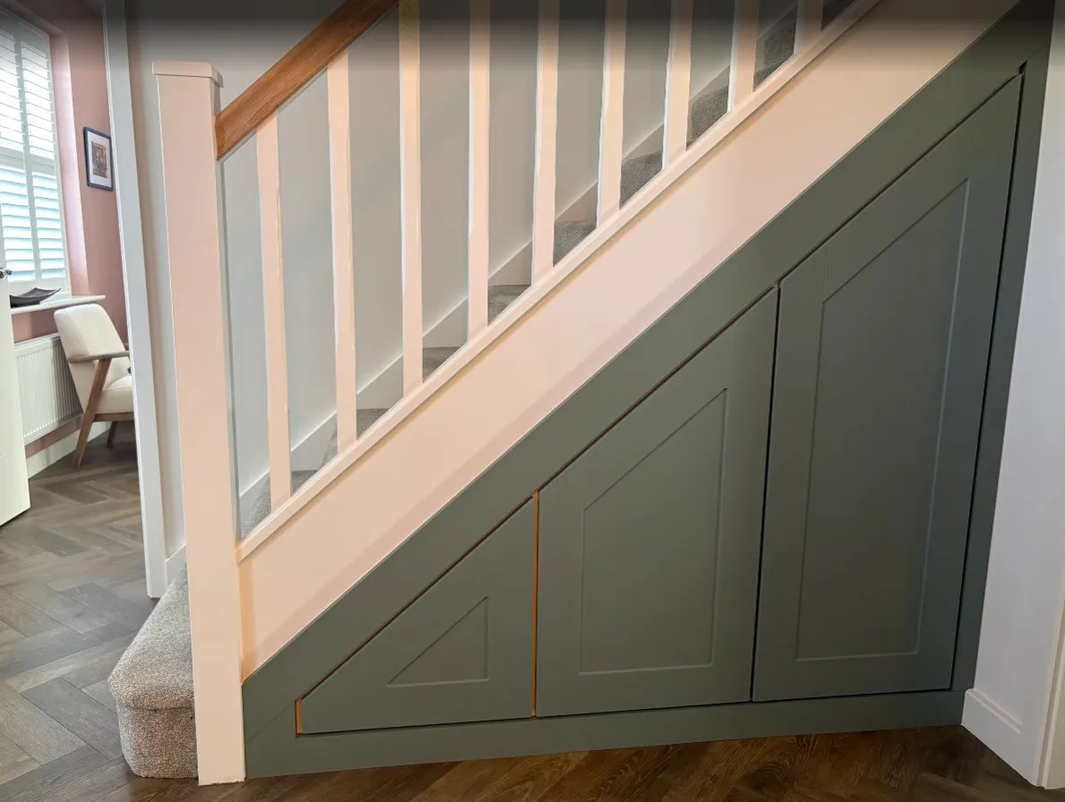 Three sage green shaker doors under a staircase with a pink balustrade