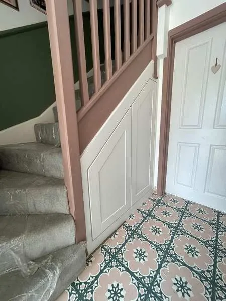 Two white doors under a stair with a pink balustrade, on a patterned tiled floor