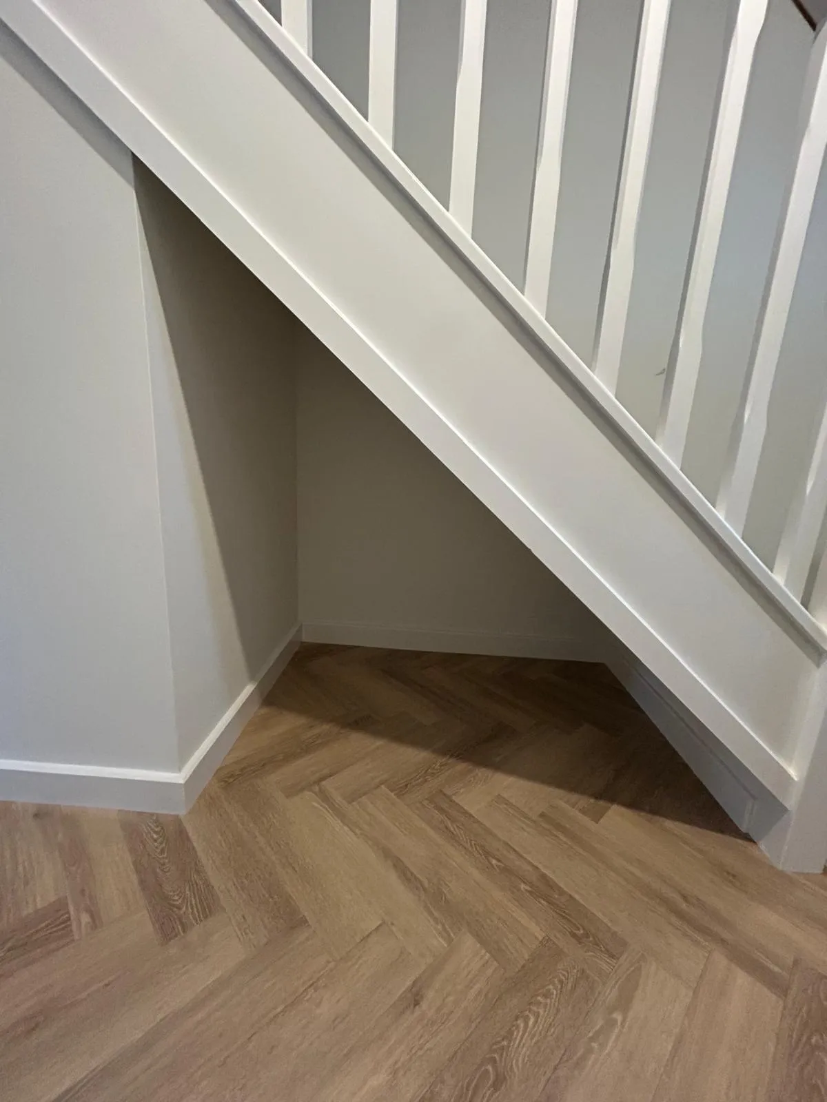 An empty open alcove beneath a white spindled staircase with a herringbone floor, before storage was fitted