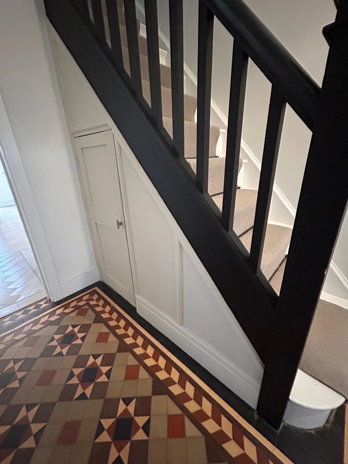 The original under stairs cupboard with a single handled door, beneath a black balustrade staircase and Victorian tiled floor