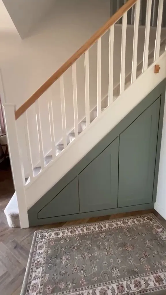 Sage green under stairs storage with three angled doors, closed, beside a white staircase with oak handrail