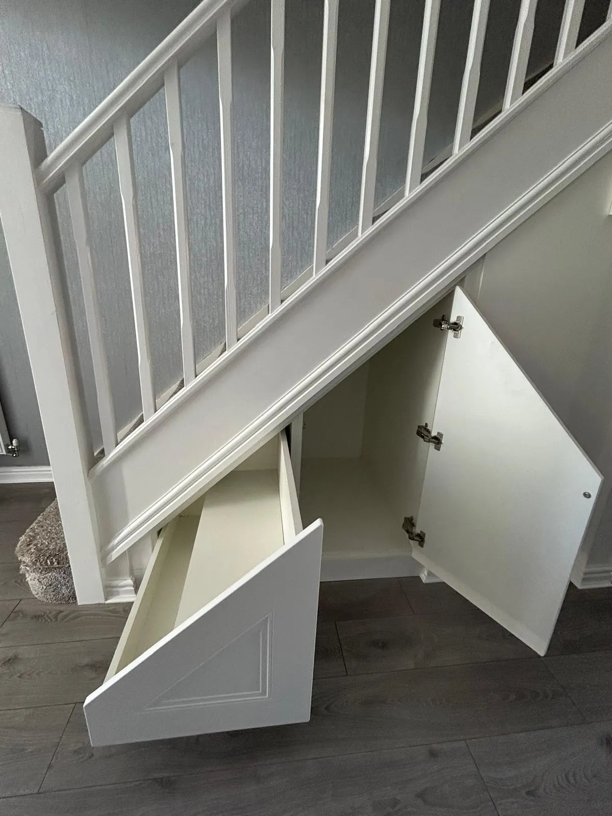 A white under stairs run with an angled drawer and a hinged cupboard door open, showing the deep bays inside